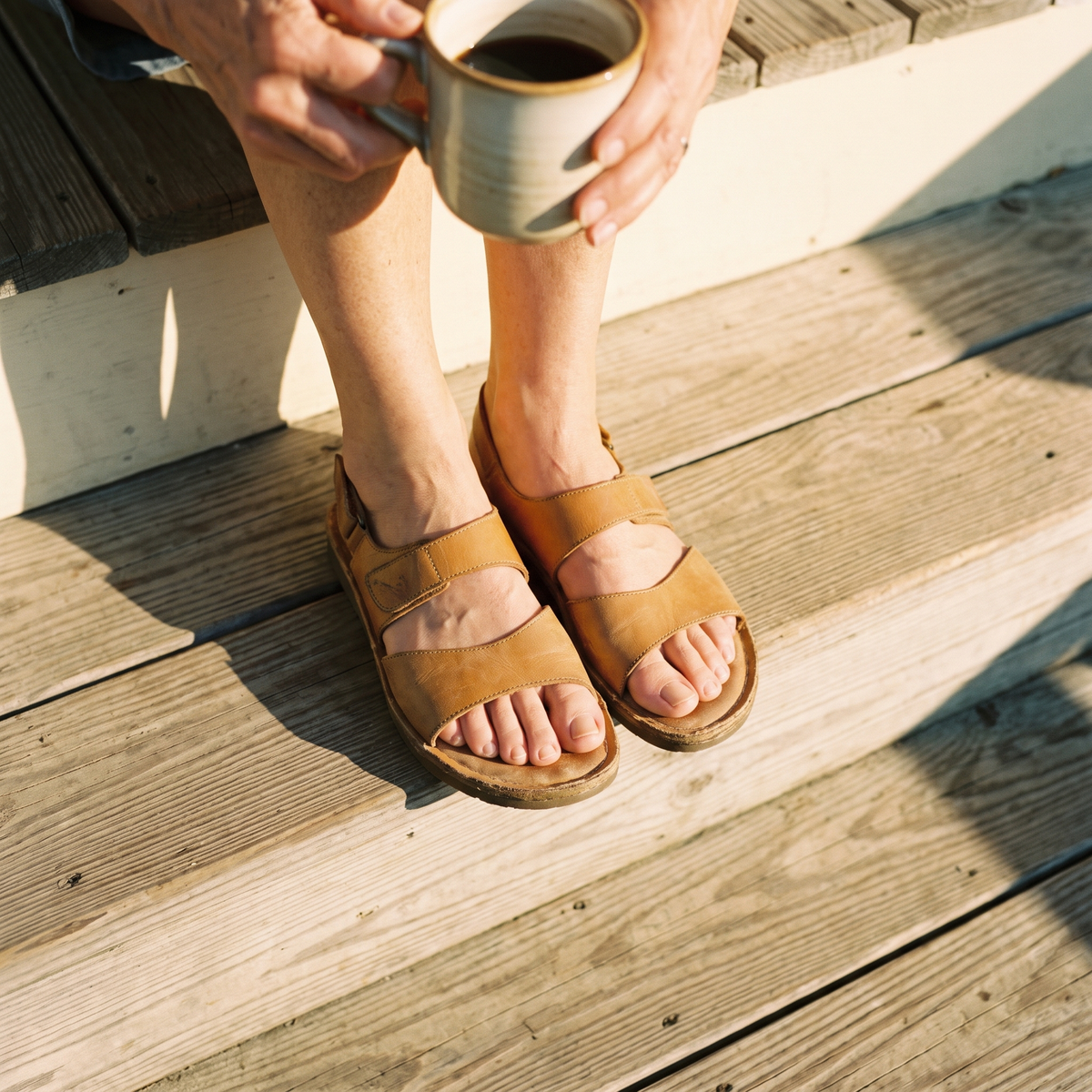 A woman in her sixties sitting on a sunlit porch step in open sandals with a cup of coffee