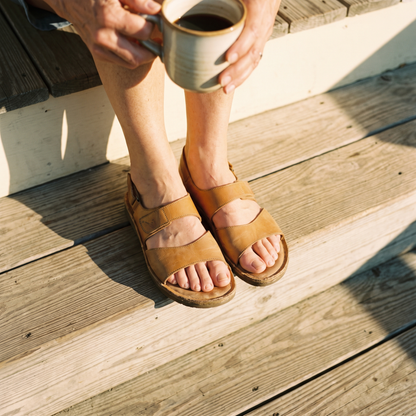 A woman in her sixties sitting on a sunlit porch step in open sandals with a cup of coffee